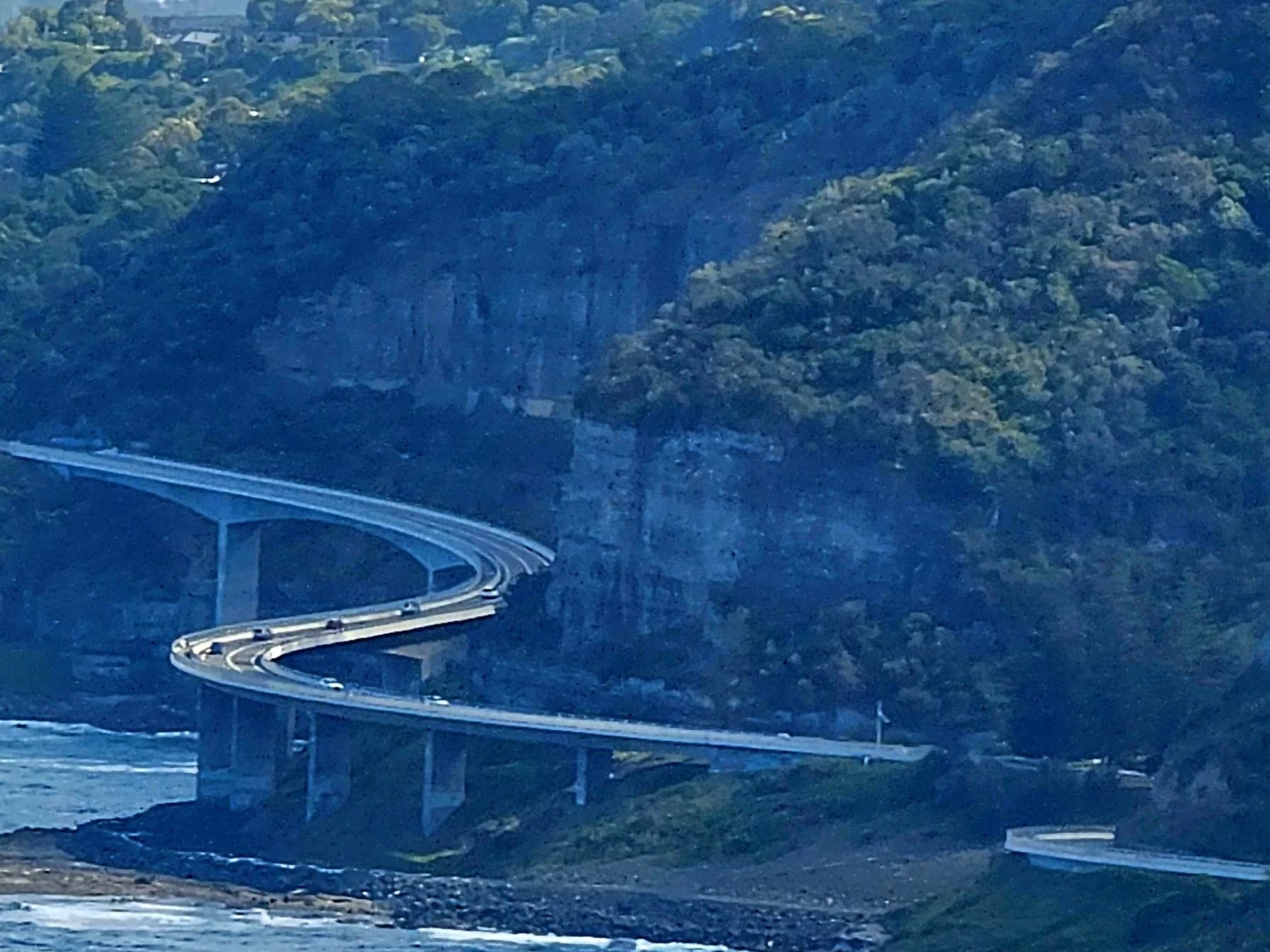 The Sea Cliff Bridge curving around the cliffs above the ocean, seen from Bald Hill