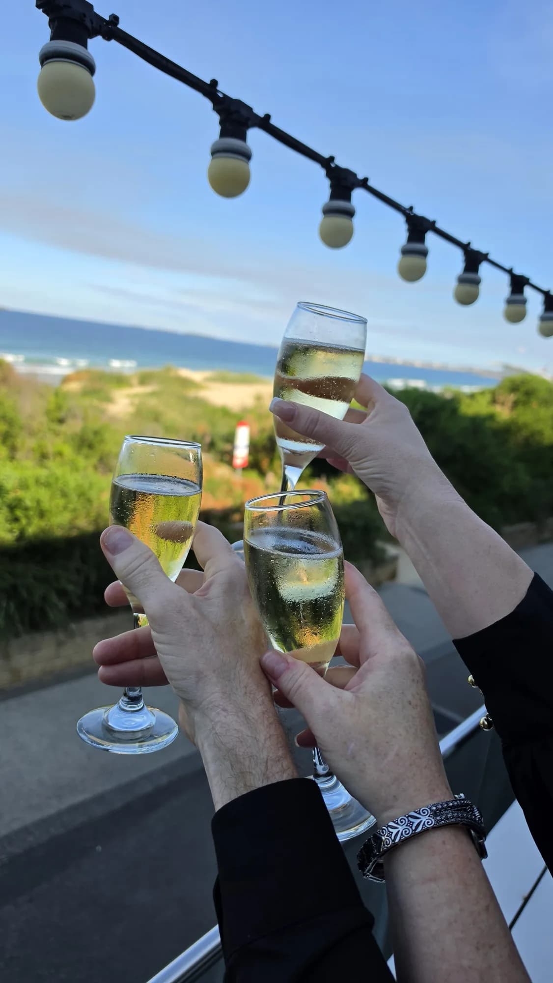 Glasses of sparkling wine raised under festoon lights, with the dunes and North Beach behind Steamers
